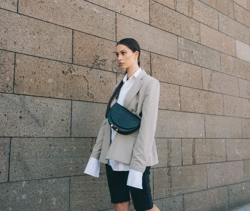 woman in beige blazer, black shorts, and pair of chunky heeled sandals walking near wall